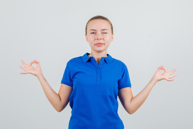 Young woman doing meditation and winking eye in blue t-shirt