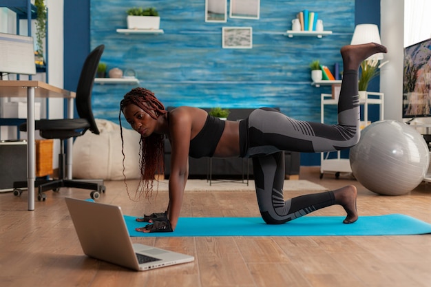Young woman doing straight leg donky kick pushing left foot up keeping leg bent, watching tutorial video on laptop screen