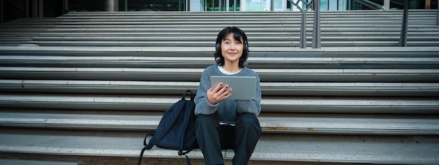Free photo young woman draws on her tablet listens to music in headphones asian girl sits on stairs and does