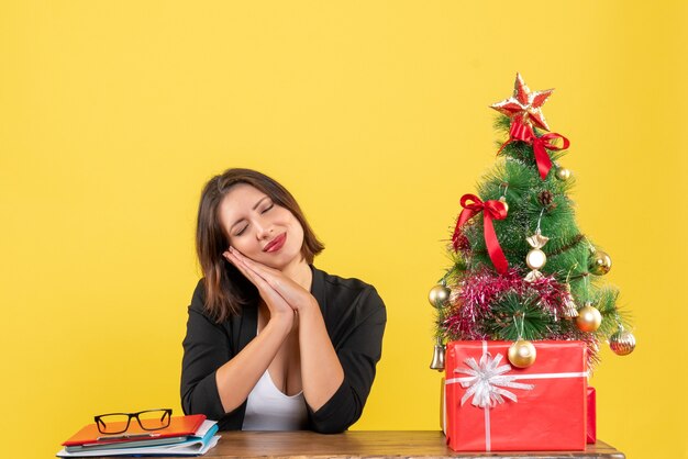 Young woman dreaming about something sitting at a table near decorated Christmas tree at office on yellow 