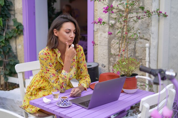 Young woman in a dress in a bright street cafe with a laptop works remotely on her own schedule from anywhere in the world online