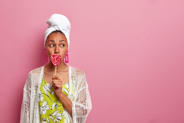 young woman dressed in casual clothing, looks seriously aside, covers mouth with sweet tasty heart shaped lollipop, isolated on pink wall, blank copy space