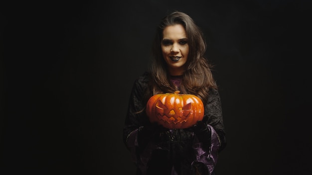 Giovane donna vestita come una strega che tiene una zucca per halloween guardando la telecamera su uno sfondo nero.