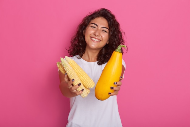 young woman dressed in white casual t shirt showing zucchini and corncob to camera, has happy facial expresion, posing with toothy smile. Raw food diet and healthy eating concept.