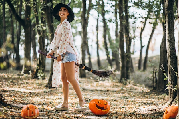 Молодая женщина, одетая в шляпу с метлой на Хэллоуин в лесу