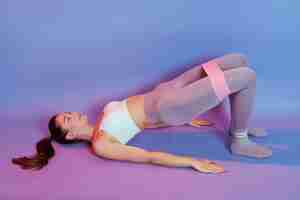 Free photo young woman dresses gray leggins and white tank top lying on floor and doing gluteal bridge with rubber resistance band, looking up, training buttocks, isolated over color background.