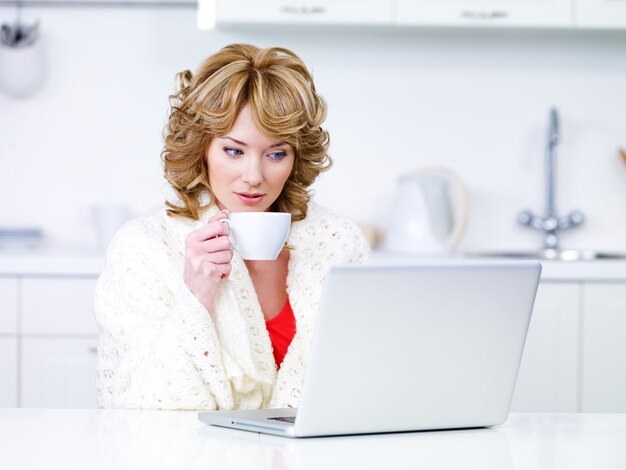 Young woman drinking coffee and using laptop in the kitchen