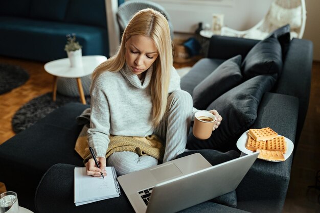 コンピューターで作業し、リビングルームでメモを取りながらコーヒーを飲む若い女性