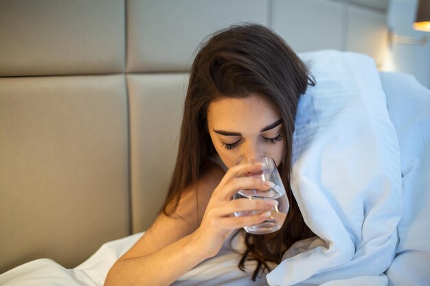 夜にベッドでコップ一杯の水を飲む若い女性寝る前にコップ一杯の水を飲む女性彼女はベッドに横たわっています