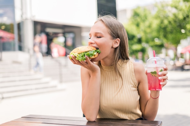 Молодая женщина пьет лимонад и гамбургер в кафе уличной еды
