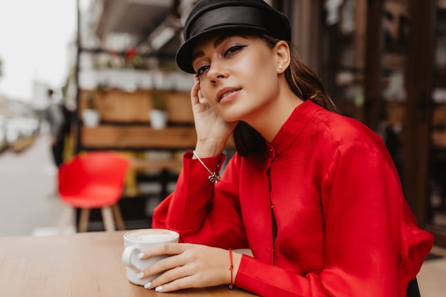 Young woman drinks coffee in street cafe. Lady with beautiful makeup in expensive blouse is mysteriously