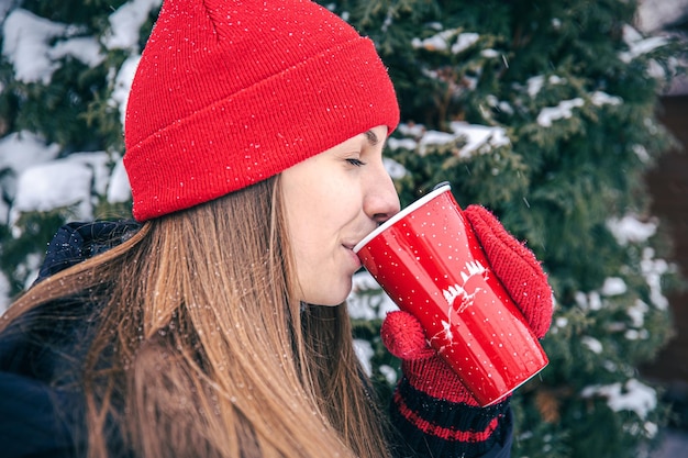 A young woman drinks a hot drink from a red thermal cup in winter