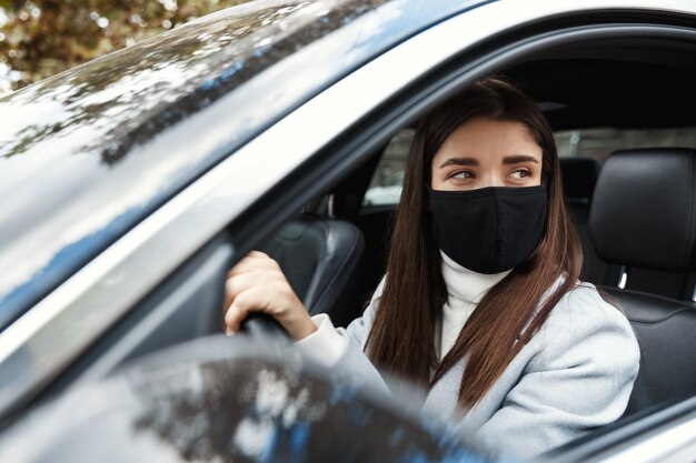 車に座って、フェイスマスクで働くために運転している若い女性ドライバー