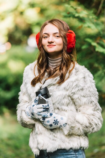 Young woman in earmuffs holding smartphone and thermos