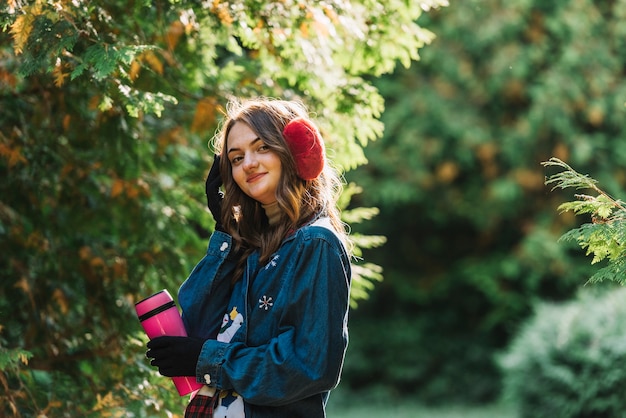 Young woman in earmuffs holding thermos near branches