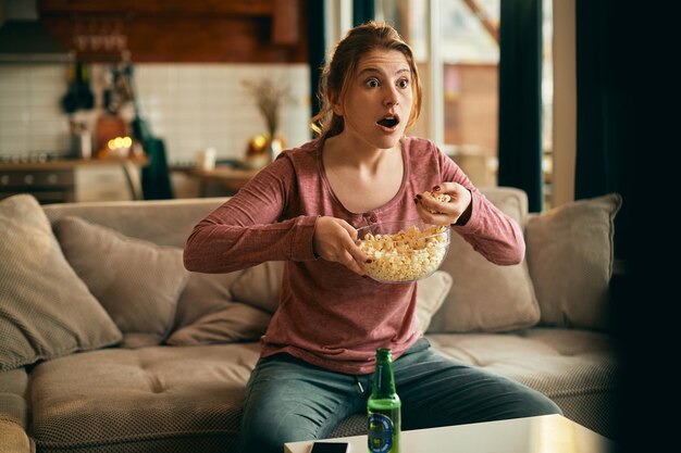 Free photo young woman eating popcorn while watching in disbelief something on a tv