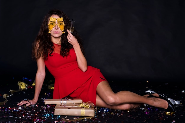 Young woman in evening dress with glass and funny mask near present boxes 