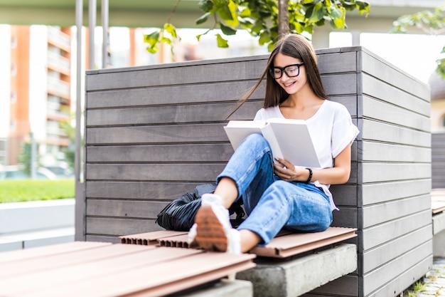 Молодая женщина в очках сидит на скамейке и читает книгу