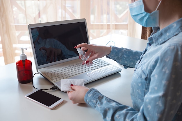 Young woman in face mask disinfecting gadgets surfaces on her workplace