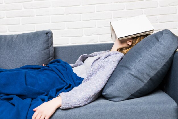 Young Woman fallen asleep while reading lying in the sofa with book on the head