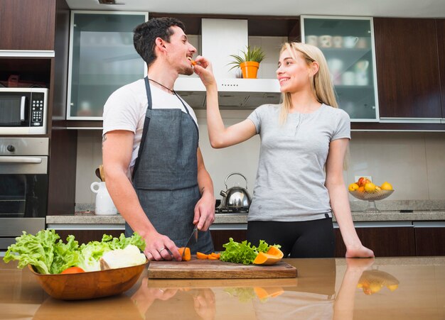 Молодая женщина кормит кусочек моркови своему мужу на кухне
