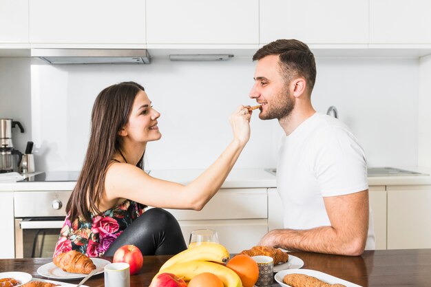 Young woman feeding her boyfriend with breakfast on the table in the kitchen