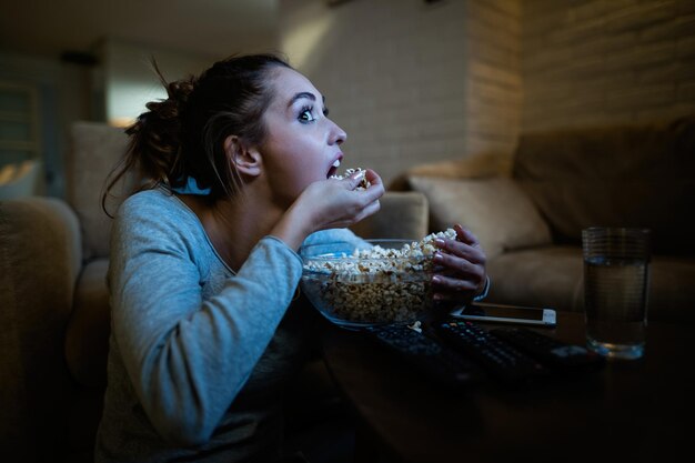 Молодая женщина чувствует голод и ест попкорн из миски во время просмотра фильма ночью дома