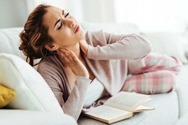 Young woman feeling pain in her neck while lying down on sofa in the living room