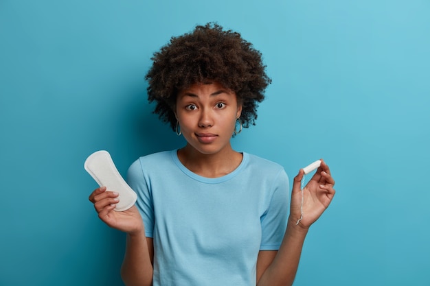 Young woman feels hesitant, chooses between sanitary pad and tampon during critical days, has good hygiene protection, regular menstruation cycle, isolated on blue wall. Women and menses