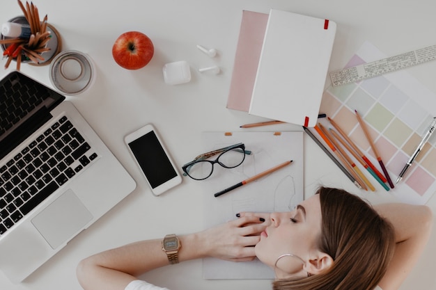 Young woman fell asleep on desktop, surrounded by documents, notes, ruler and devices