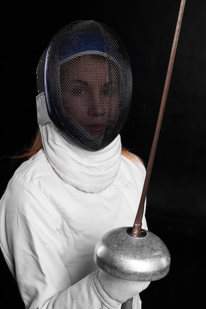 Free photo young woman in fencing mask and white costume keeps sword and look to the front