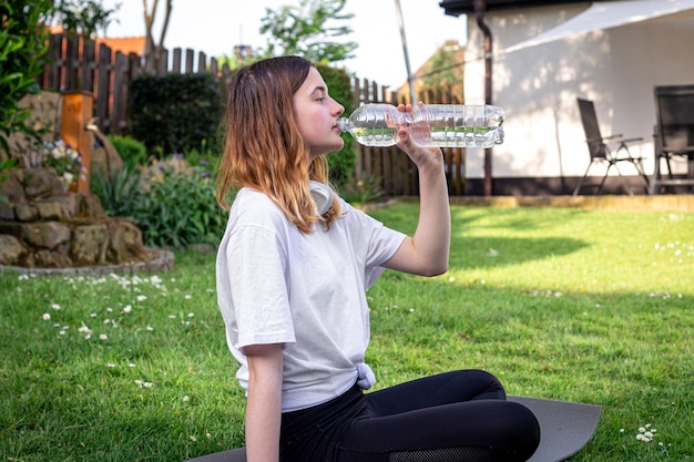 A young woman on fitness mat drinks water in nature sports concept