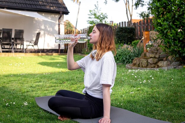 A young woman on fitness mat drinks water in nature sports concept