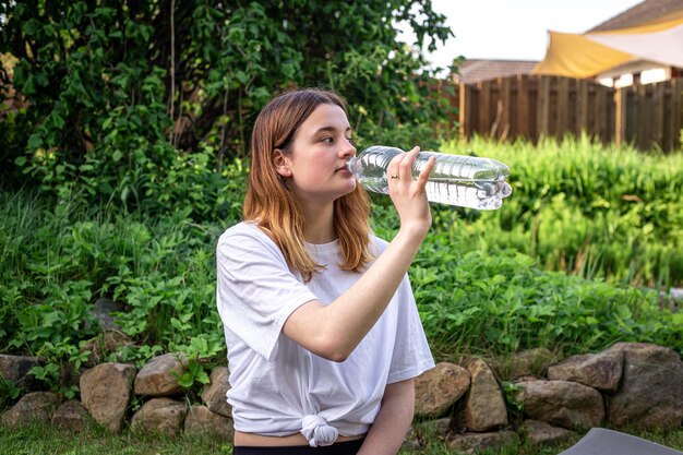 A young woman on fitness mat drinks water in nature sports concept