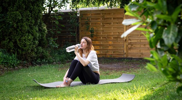 A young woman on fitness mat drinks water in nature sports concept