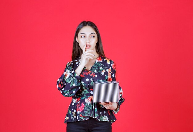 Young woman in floral shirt holding a silver gift box and looks thoughtful