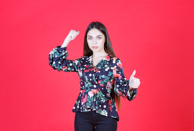 Giovane donna in camicia floreale in piedi sul muro rosso e dimostrando i suoi muscoli del braccio