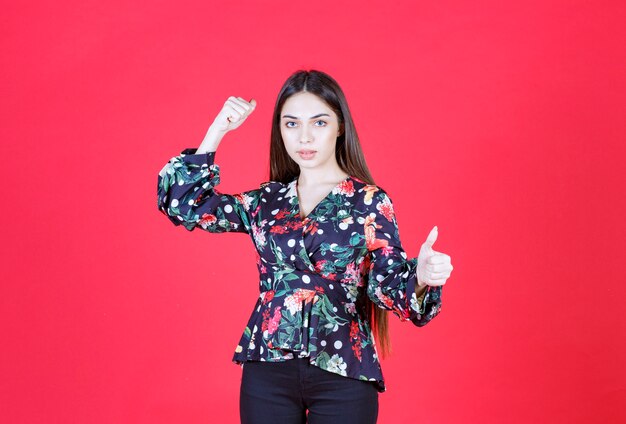 Young woman in floral shirt standing on red wall and demonstrating her arm muscles