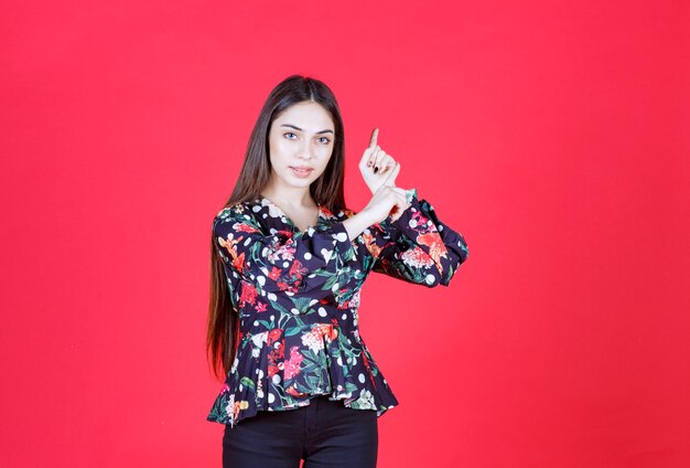빨간 벽에 서서 거꾸로 보여주는 꽃무늬 셔츠를 입은 젊은 여자