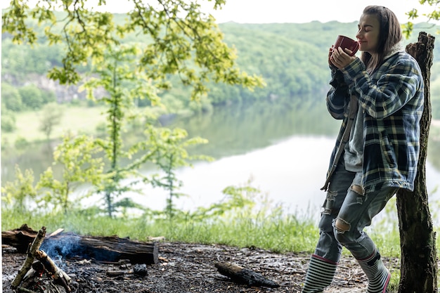 A young woman in the forest near the river warms up by the fire and drinks a hot drink.