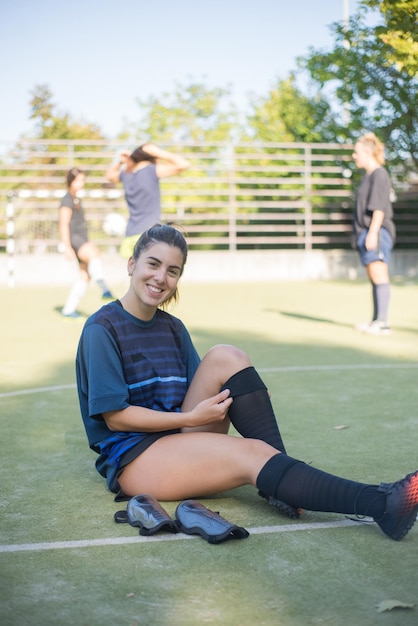 Молодая женщина одевается для тренировки. Женщина-игрок сидит на траве, смотрит в камеру, улыбается. Концепция футбола, спорта, досуга
