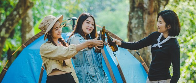 텐트의 캠프 의자에서 휴식을 취하는 젊은 여성과 여자 친구 여행자들은 함께 즐겁고 행복한 이야기를 나누며 캠핑을 하며 맥주를 마시고 있다