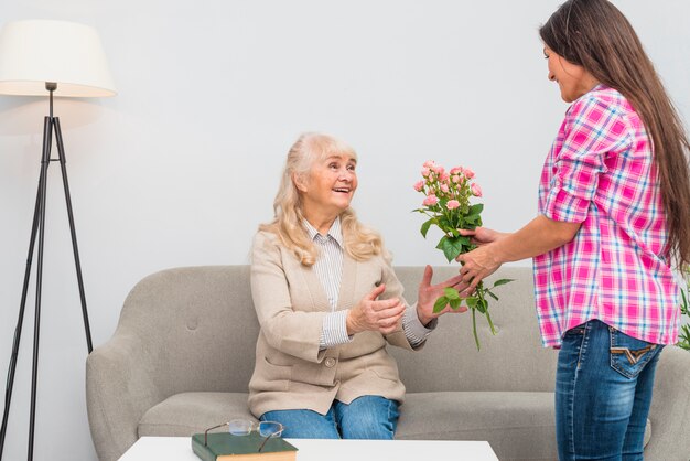 Young woman giving rose bouquet to her senior mother sitting on sofa at home