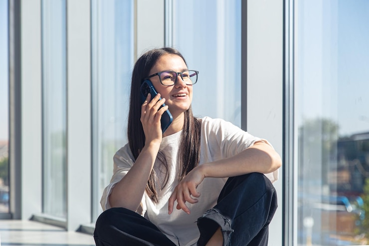 無料Photo メガネをかけた若い女性が窓の近くでスマートフォンで話している