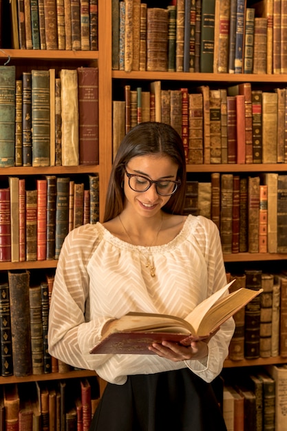 Young woman in glasses reading book near shelf