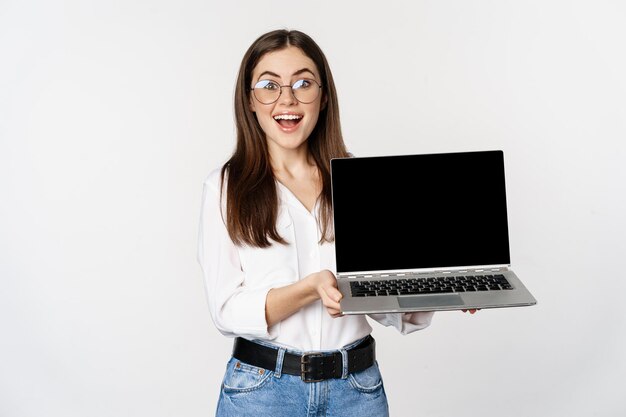 Young woman in glasses showing laptop screen, demonstrating promo on computer, website or store, standing over white background.