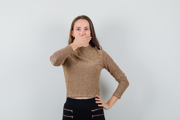 Young woman in gold gilded sweater and black pants covering mouth with hand and holding hand on waist and looking surprised , front view.