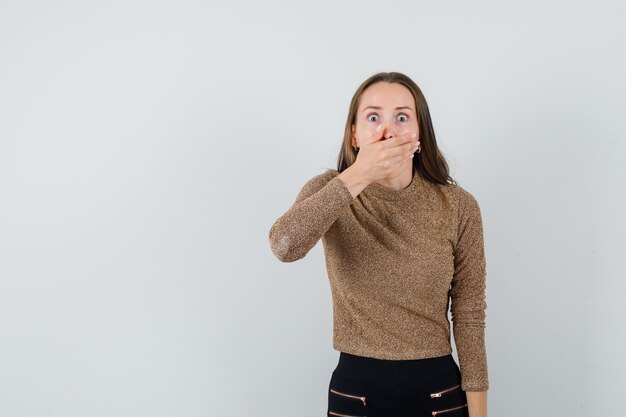 Young woman in gold gilded sweater and black pants covering mouth with hand and looking surprised , front view.