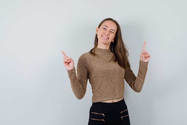 Young woman in gold gilded sweater and black pants pointing up with both index fingers and winking and looking happy