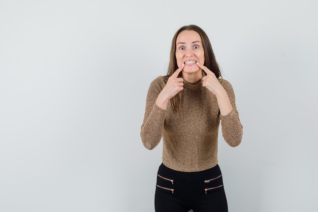 Young woman in gold gilded sweater and black pants putting index fingers near mouth, forcing smile and looking happy , front view.